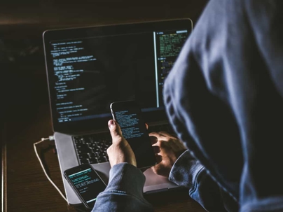 Cyber criminal wearing a navy blue hoodie sits in a dark room looking at code on a smartphone and entering it into a laptop