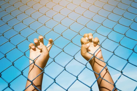 Close up view of a child's hands gripping chain link fencing with blue skies in the background